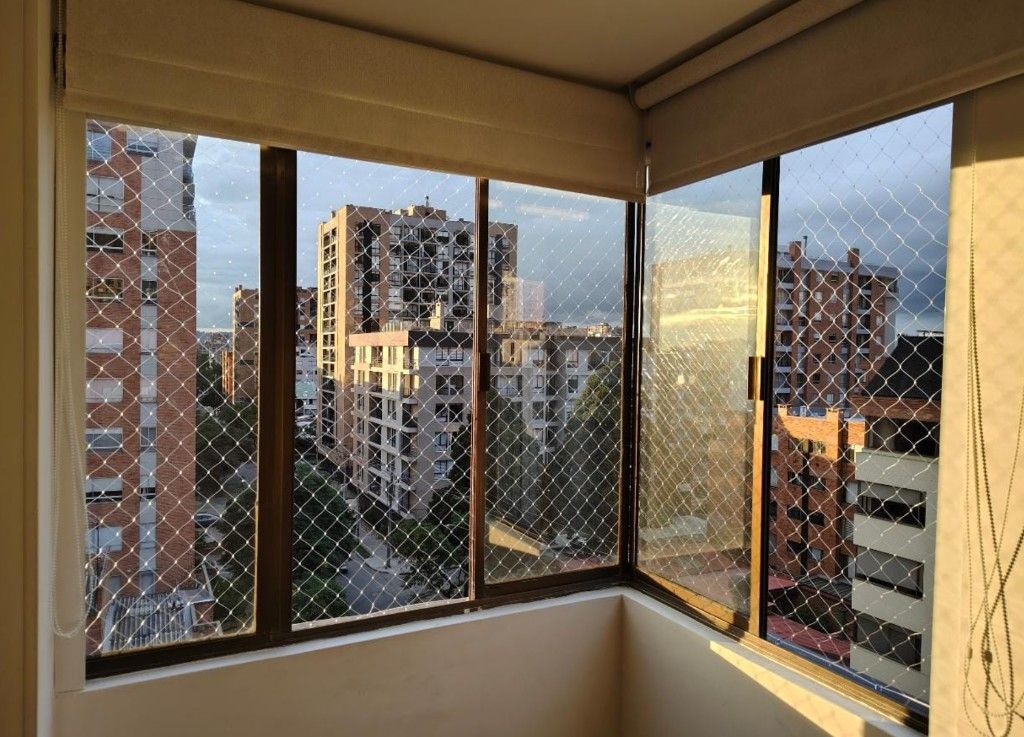Vista interior de ventanas de departamento con malla romboidal y edificios al fondo