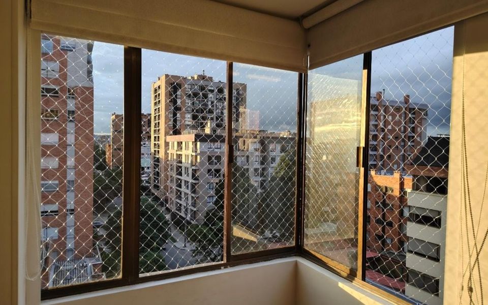 Vista interior de ventanas de departamento con malla romboidal y edificios al fondo